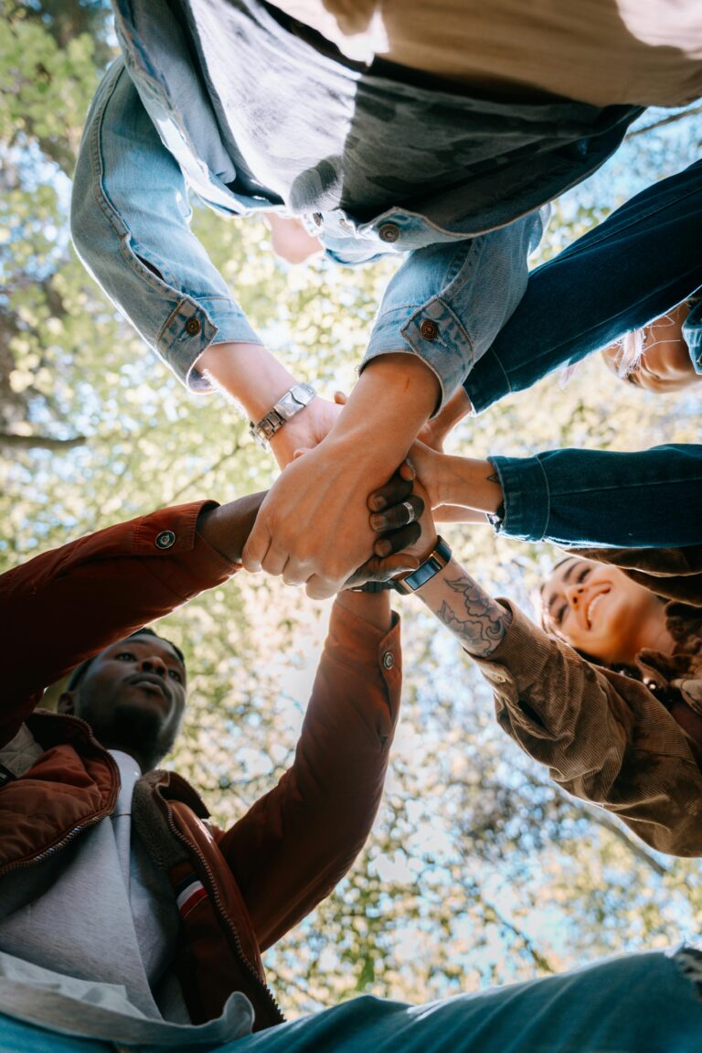 A diverse group of adults joining hands outdoors, symbolizing unity and teamwork from a low angle perspective.